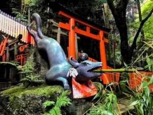 Estatua de zorro kitsune con bambú en el santuario Fushimi Inari de Kioto, rodeada de vegetación verde intensa durante un viaje de verano a Japón.